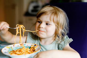 Adorable toddler girl eat pasta spaghetti with tomato bolognese with minced meat. Happy preschool child eating fresh cooked healthy meal with noodles and vegetables at home, indoors.