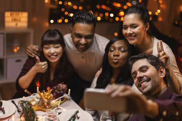 Poster - holidays, party and celebration concept - multiethnic group of happy friends having christmas dinner at home and taking selfie with smartphone