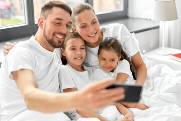 Poster - family and people concept - happy mother, father and two daughters taking selfie with smartphone in bed at home