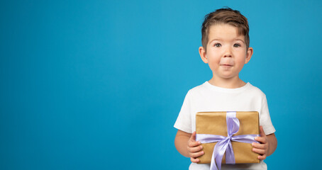 Caucasian little boy in white t-shirt holding gift box on blue background. Holiday concept