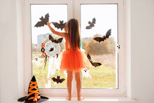 The Girl Decorates The Window In The Room With Bats And Halloween Ghosts Standing On The Windowsill. Preparing For A Masquerade Party. A Girl In An Orange Witch Dress.