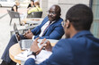 © xartproduction - Two dark-skinned African American businessmen in suits and glasses with briefcases sit at a table in an outdoor city cafe.