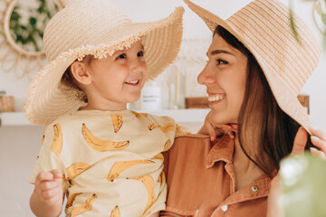 A happy young woman holds her daughter in her arms, they laugh and try on straw hats.Bright cozy home interior with indoor plants.Family, vacation, summer concept.