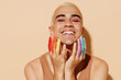 Young smiling satisfied cool happy fun blond latin gay man with make up fingers painted in rainbow flag colors wear beige tank shirt hold face isolated on plain light ocher background studio portrait.