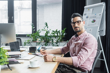 Wall Mural - Muslim businessman looking at camera near cup of coffee, laptop and notebook in office