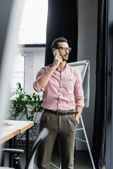 Wall Mural - Arabian businessman holding hand in pocket of pants while talking on smartphone in office