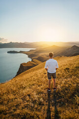 Wall Mural - A young man travels along the mountain coast at sunset. Rear view A man stands on a mountainside and enjoys the view. The concept of freedom and achievement of goals.