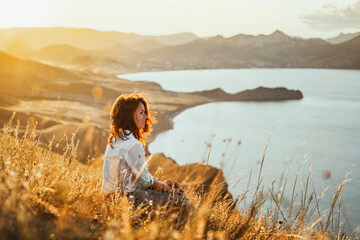 A beautiful young woman tourist is sitting on top of a mountain and enjoying the view of the sunset sea and mountains. The concept of freedom and meditation, an active lifestyle.