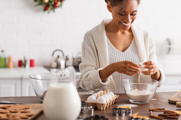 Canvas Print - Happy african american woman pouring egg in flour near blurred cookies and cinnamon sticks