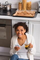 Canvas Print - Cheerful african american woman with tea and smartphone sitting near baked christmas cookies in kitchen