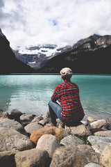 Wall Mural - Woman in red plaid shirt sitting on rocks admiring the view of lake and mountains