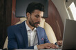 © Oleksii - Handsome Arabian businessman using laptop computer, typing, working online sitting in airplane. Confident middle eastern entrepreneur flying luxury private jet. Successful business concept