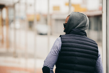 Rearview of a guy with a hood wearing a shirt, vest jacket, and sunglasses