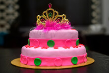 Two-tier Fondant Cake Design With A Tiara On Top, Decorated With Icing Flowers. Delicious Pink-red Color-themed Round Cake On Yellow Cake Tray Against The Dark Background.