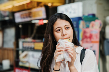 Wall Mural - hungry charming asian japanese girl trying taiwanese local street food in plastic bag and looking aside under sunshine. beautiful lady tourist enjoy traditional snack outdoor market in summer time