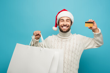 cheerful man in knitted sweater and santa hat showing credit card and shopping bags isolated on blue