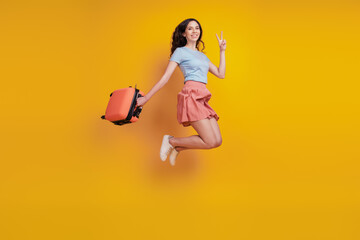 Full length photo of young woman happy positive smile bag suitcase jump show peace v-sign isolated over yellow color background