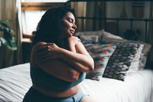 Curly Haired Overweight Young Woman In Blue Top And Shorts With Satisfaction On Face Accepts Curvy Body Shape In Stylish Bedroom.