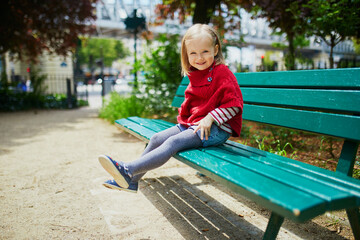 Wall Mural - Happy cheerful toddler girl sitting on the bench on a street of Paris