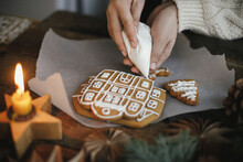 Decorating Christmas Gingerbread Cookies With Icing On Wooden Table With Candle And Ornaments. Close Up Of Making Gingerbread House With Frosting. Atmospheric Moody Image. Holiday Preparations