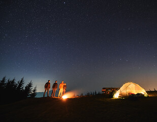 Wall Mural - Four travelers warming by warmth of bonfire on cool night on mountain hill. White lighted tent, wooden table on green grass in tourist camp. Starry sky over the mountains. Concept of night camping.