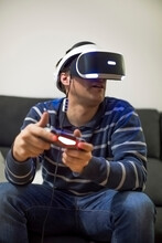 Young Man Wearing VR Glasses Sitting On Couch At Home Playing Video Game