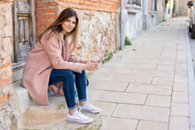 Portrait Of A Young Woman Sitting At House Entrance Holding Cell Phone