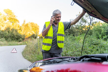 Senior Man Standing At His Broken Car Wearing A Safety Vest And Using His Smartphone