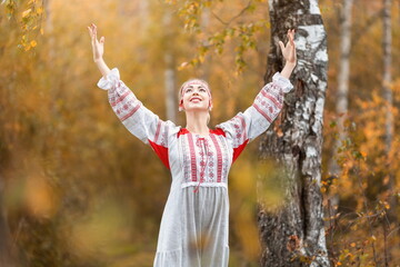 Wall Mural - Portrait of young beautiful smiling woman in folk traditional slavic clothes waving hands  and asking for blessing from spirits at nature in autumn forest. Pagan slavs rituals and traditions concept