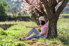 Happy Teenage Girl Leaning Against Almond Tree Looking At Twig Of Almond Blossoms