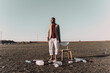 © Eloisa Ramos/Westend61 - Young man standing next to chair surrounded by plastic bottles in barren land