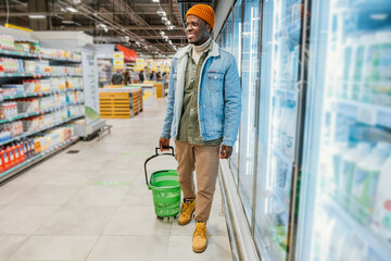 Wall Mural - Smiling African-American man with shopping basket stands between rack and refrigerators in modern supermarket department