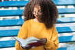 © NOVELLIMAGE/Westend61 - Smiling young woman reading book while sitting in bleachers
