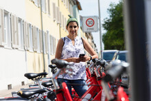 Smiling Woman Holding Smart Phone While Standing By Rental Bicycles In City