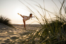 Flexible Young Woman Practicing Yoga On Sand At Beach During Sunset