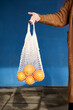 © Veam/Westend61 - Woman holding oranges in reusable bag against wall on footpath