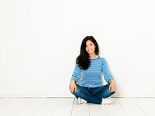 Beautiful Young Woman With Black Hair And Blue White Striped Sweater Sitting On The Ground In Front Of White Background