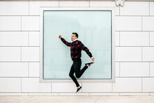 Young Man With Casual Clothes Jumping With A White Wall And A Large Window In The Background