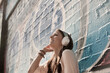 Happy young woman listening music through headphones while standing against wall with graffiti on sunny day
