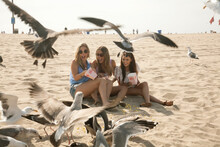 Young Female Friends Feeding Popcorn To Seagulls At Beach