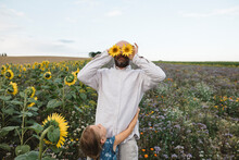 Playful Man Covering His Eyes With Sunflowers In A Field With Daughter Embracing Him