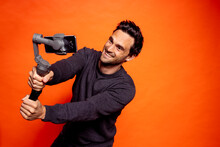 Playful Handsome Man Taking Selfie Through Smart Phone On Gimbal Against Orange Background