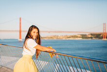 Smiling Beautiful Woman With Long Hair Standing By Railing Over River In City At Sunset