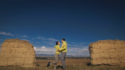 Man and woman in yellow green sportswear. Lovely couple of travelers hug and kiss near old stone enjoying highland landscape. Two travelers are walking against the backdrop of snow-capped mountains.