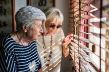 Senior Female Friends Looking Through Window Blinds At Home