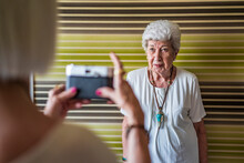 Senior Woman Photographing Female Friend Standing Against Wall At Home