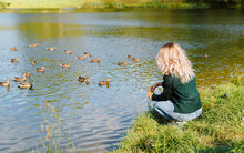 Side View Of Young Woman In Casual Clothes Sitting On Shore Near Water And Feeding Ducks In Nature. Selective Focus On Blonde Girl Throwing Food To Wild Birds Floating On Lake