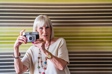 Stylish Senior Woman Photographing With Camera While Standing Against Wall At Home