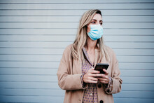 Beautiful Blond Woman Wearing Protective Face Mask While Holding Mobile Phone Against Wall