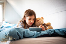 Portrait Of Girl Lying On Bed With Teddy Bear Using Digital Tablet